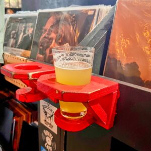A glass of hazy IPA in a rustic, red-painted wooden cupholder in a record store. The holder is mounted to a black bin of vinyl records with album art visible.